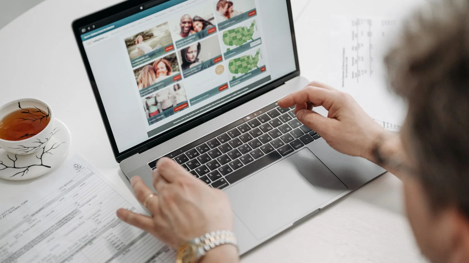A person browsing a website on a laptop at a table with tea and documents nearby.
