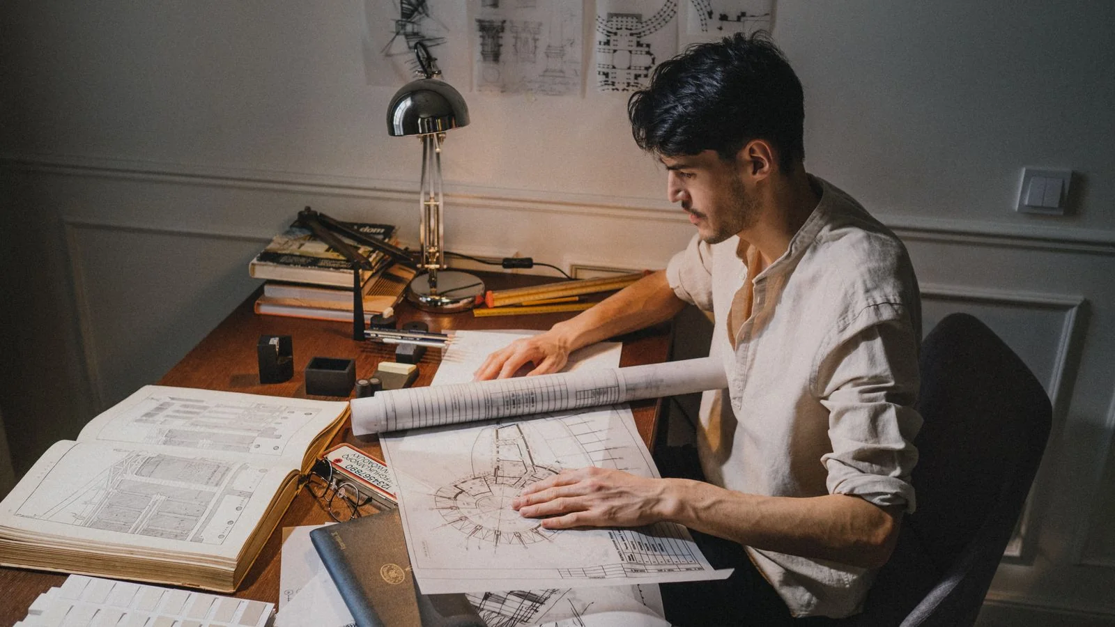 A young architect at a desk working on blueprint designs under lamp light, drafting the questions a civil engineer will be asked to answer.