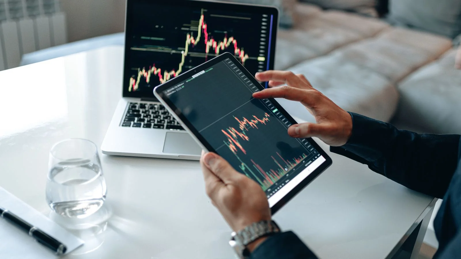 Hand scrolling market data on a tablet beside a laptop, researching a financial question