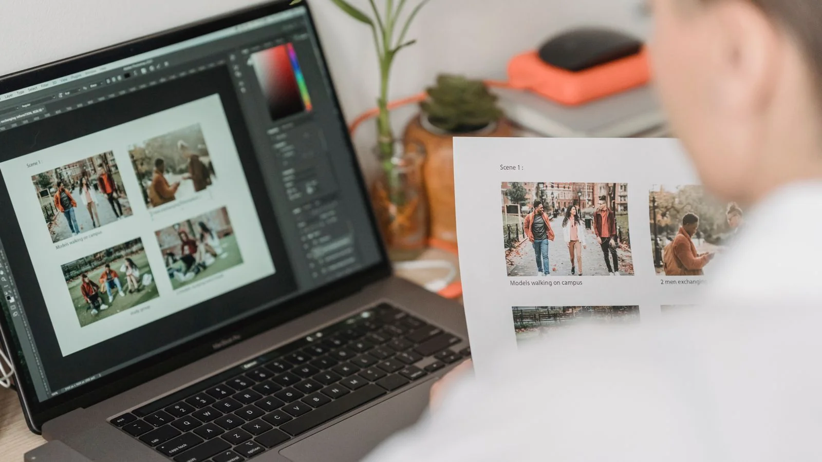 A designer at a laptop reviewing printed proofs spread across a work table.