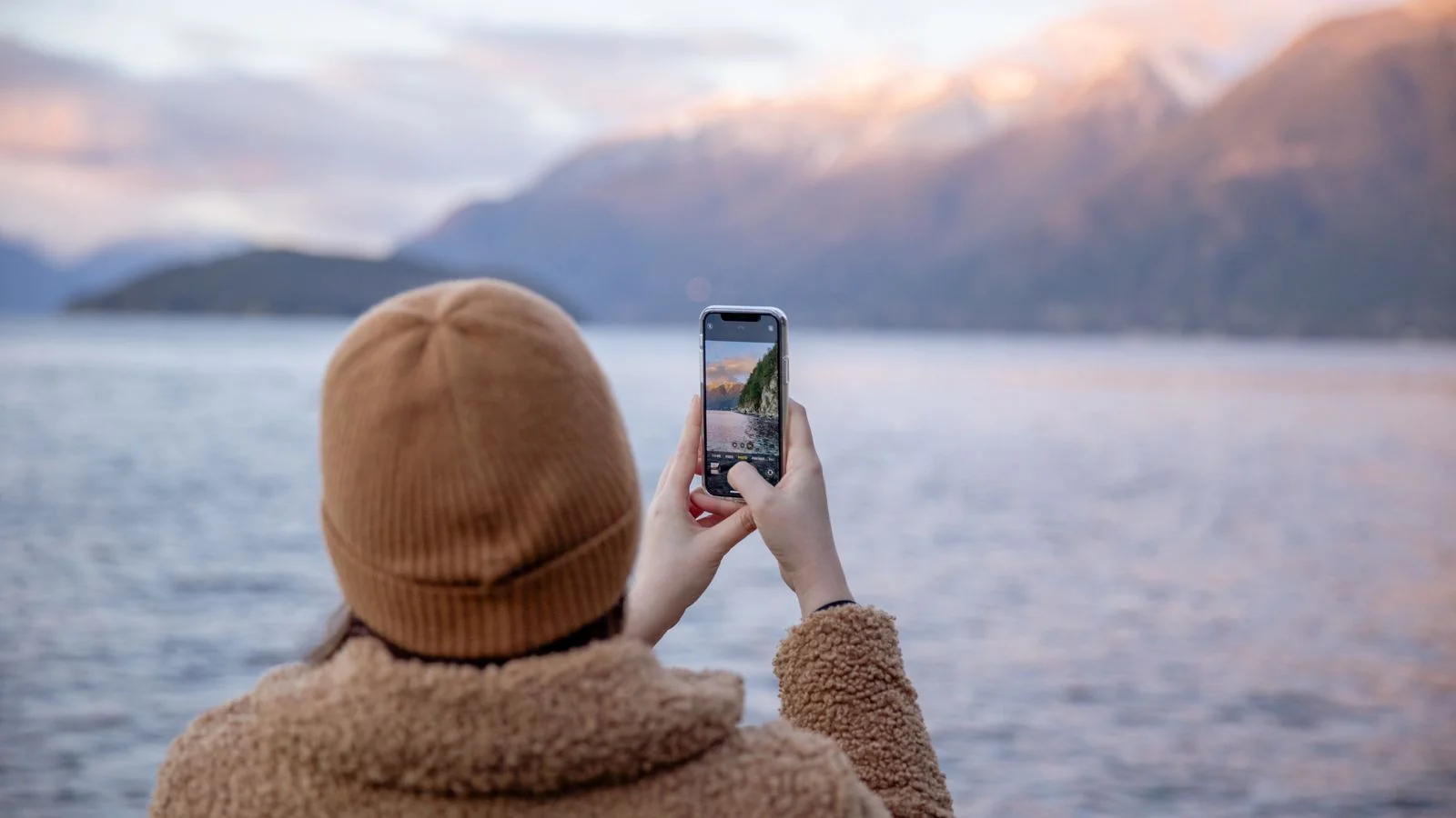 A traveler standing at a lakeside taking a photo of mountains across the water