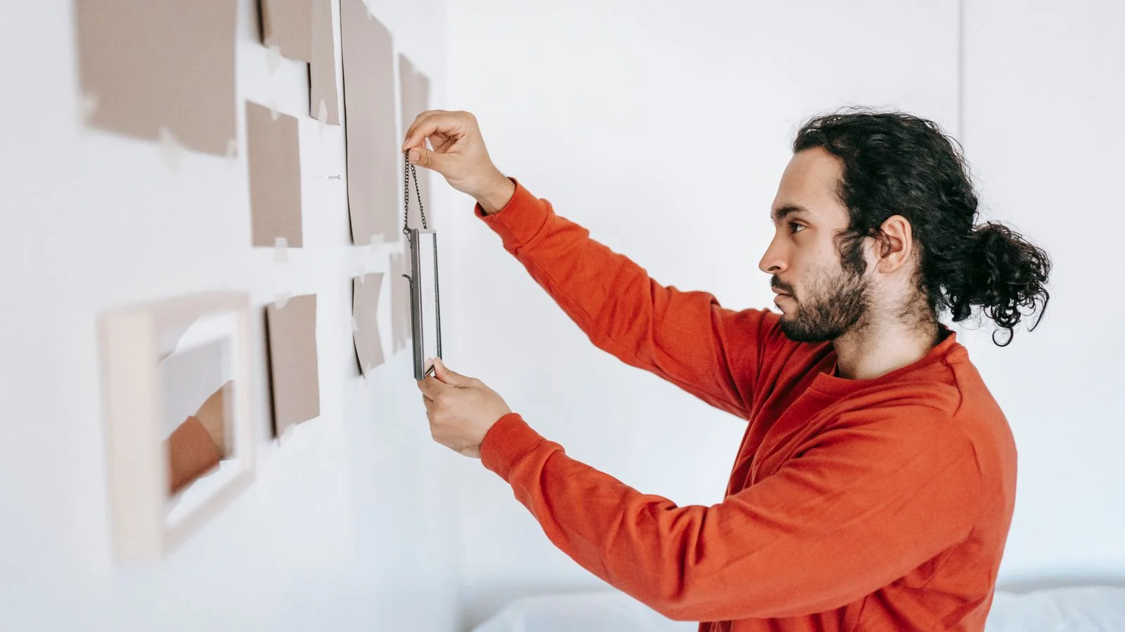 A man in a red sweater hanging framed art on a wall beside tall windows.