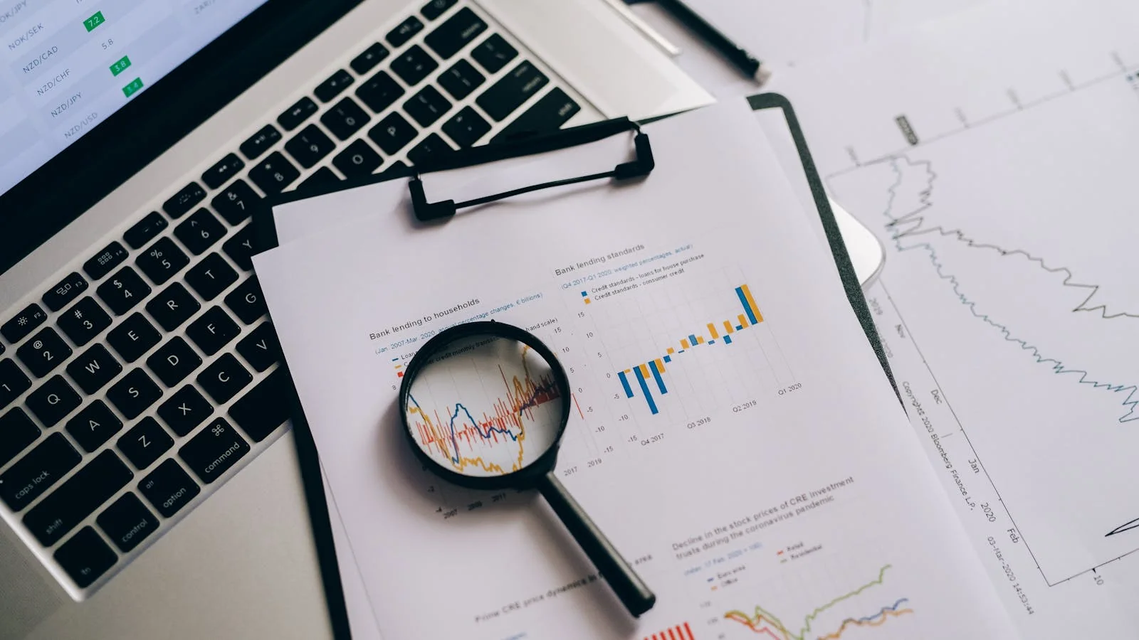 Overhead view of financial charts, a laptop screen, and a magnifying glass on a wooden desk, set up for a monthly client review.