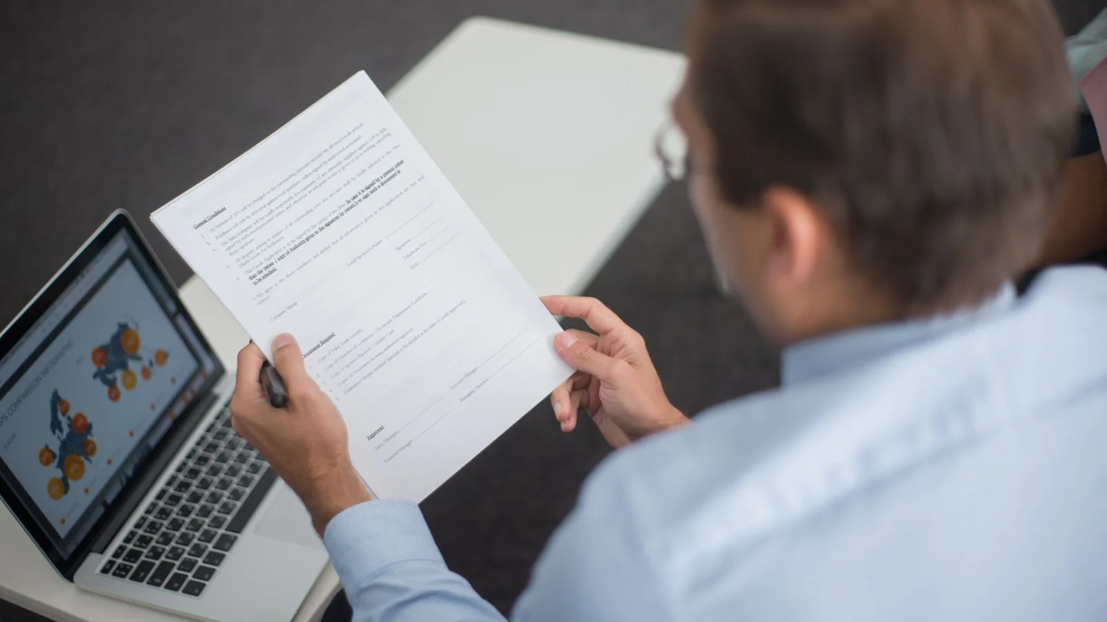 A professional in a blue shirt reviewing documents beside a laptop showing charts and search data