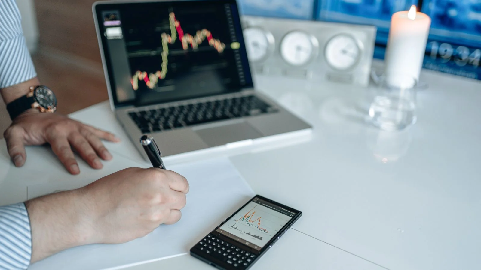 An analyst writing notes beside a laptop and phone showing trend graphs