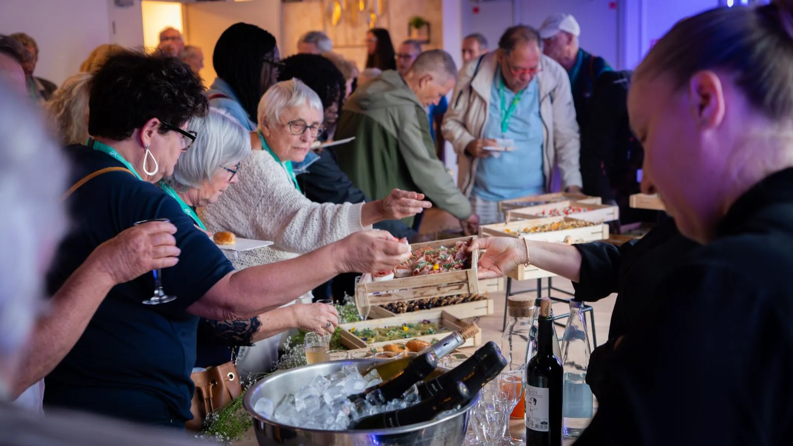 Lively crowd gathers around a buffet table at an indoor social event, like an exhibition opening reception