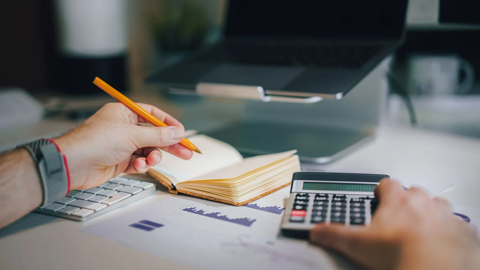 Hands working a calculator beside handwritten notes and a laptop during budget review