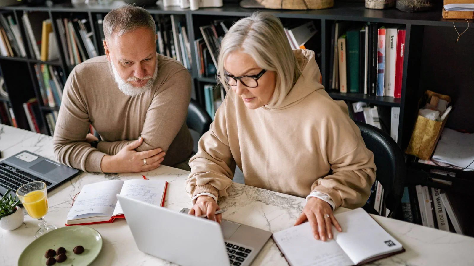 An older couple sits together using laptops at a table, researching a home project.