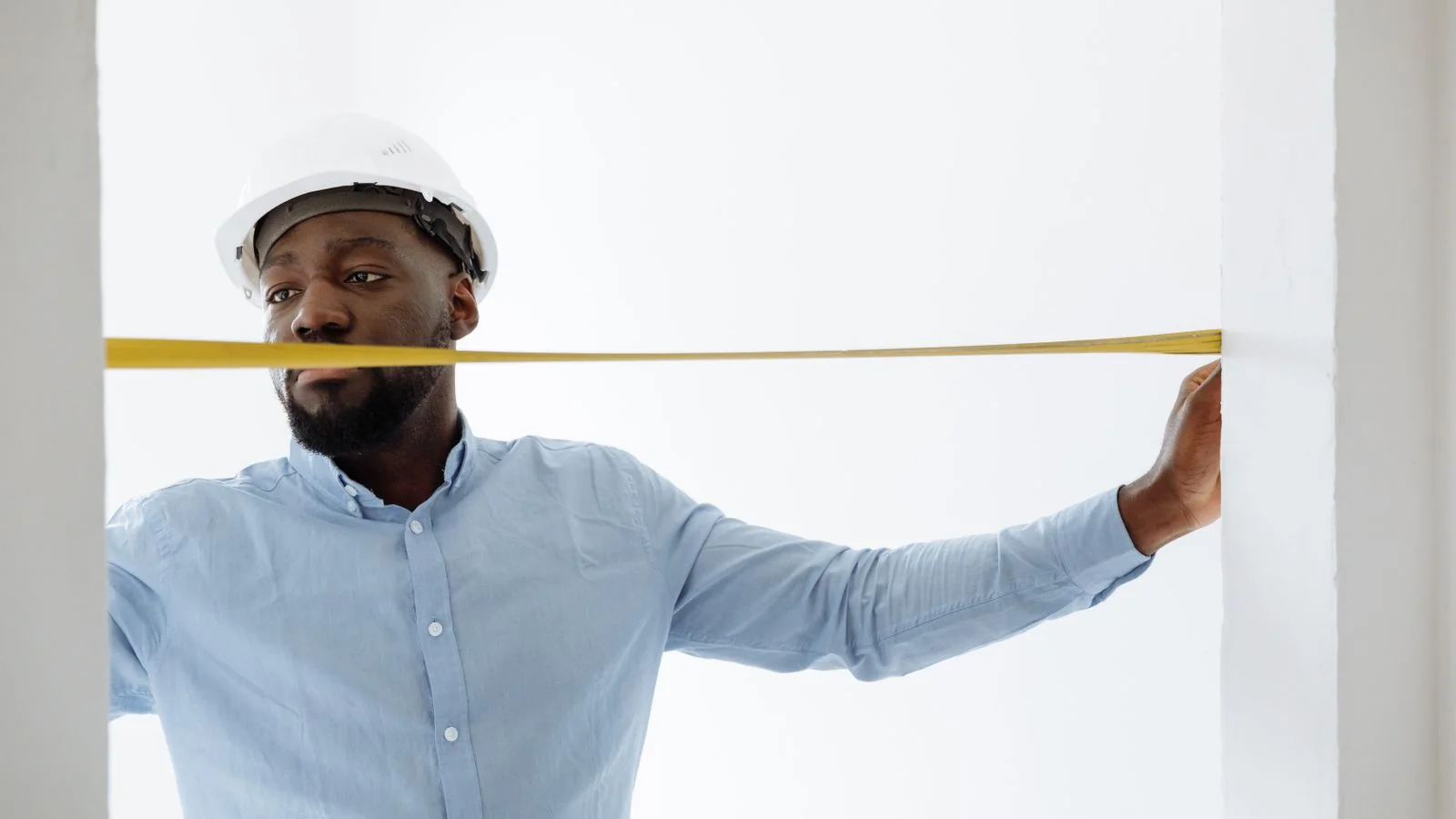 A construction worker holding a tape measure inside an unfinished room, the unglamorous middle of a project that never makes the highlight reel.