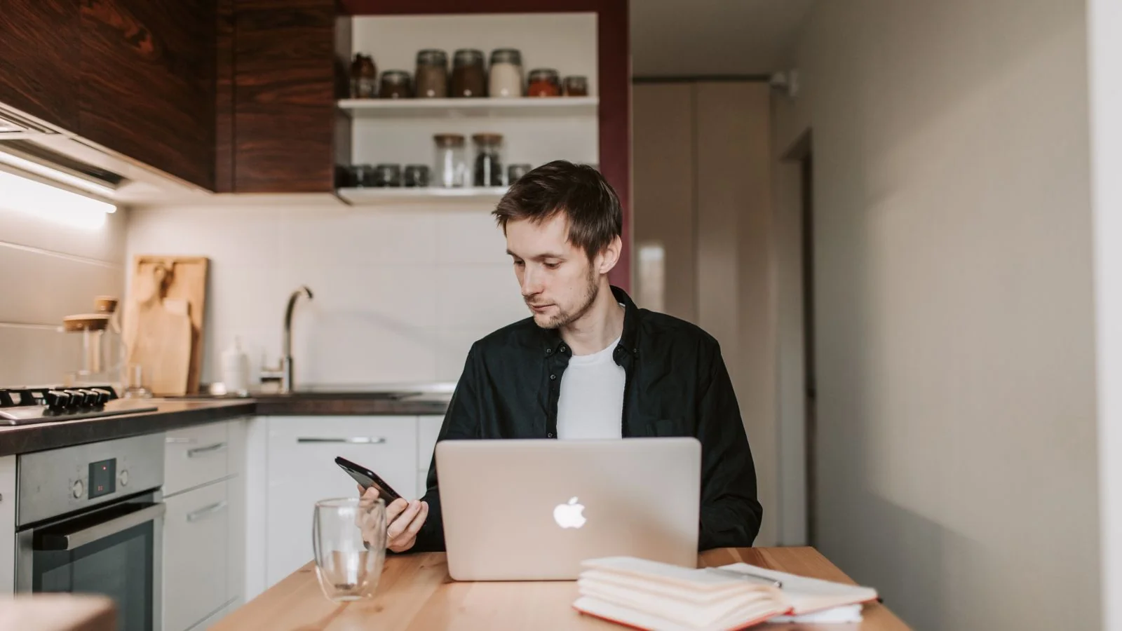 A person sits at a kitchen table using a laptop and phone to research at home.