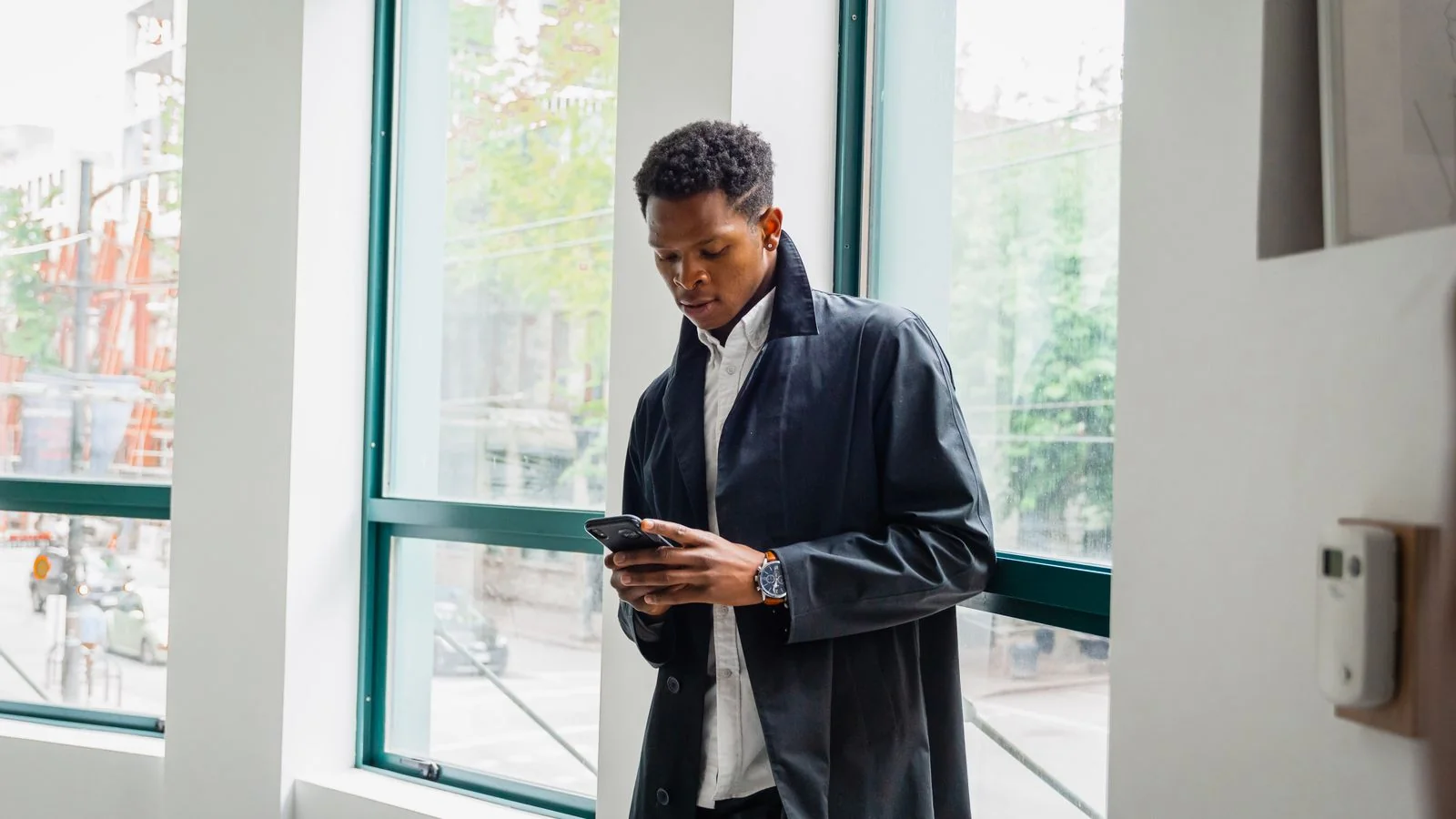 Man in a dark jacket checking his phone by an office window, the search that happens before anyone calls a hauler.