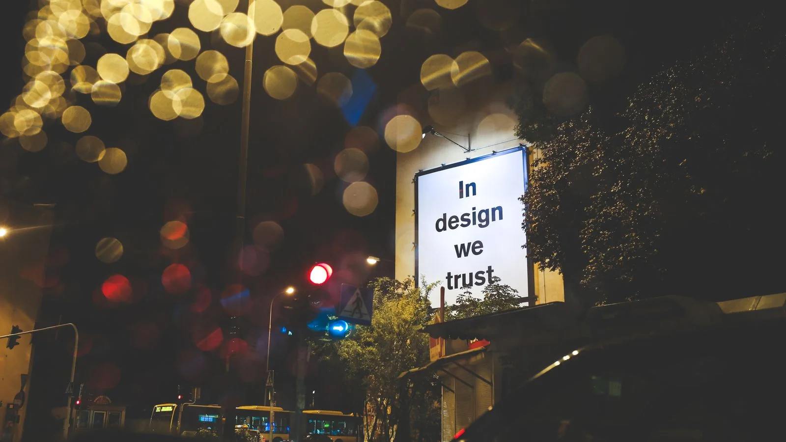 A city street at night with an illuminated billboard reading 'In design we trust,' attention rented by the day