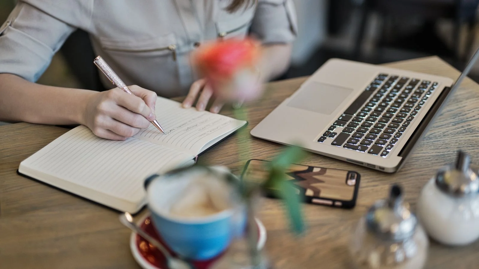 A person writing in a notebook beside a laptop, the demand-creation work of content that teaches before it sells