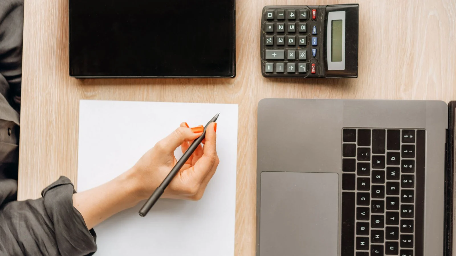 Person writing on paper beside a laptop, tablet, and calculator, pricing a writing job
