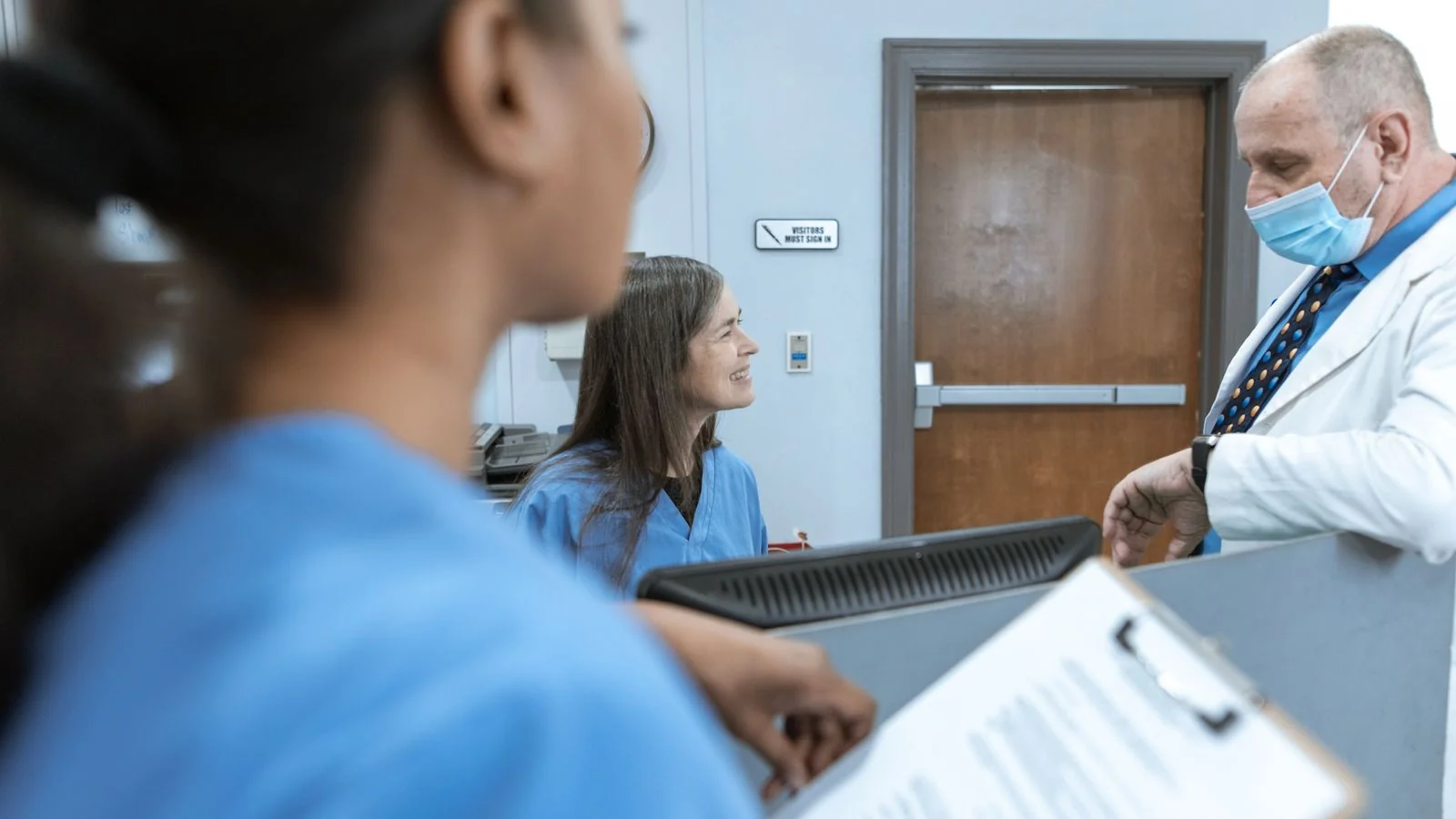 Clinicians talking at a hospital reception desk, where the practice name patients repeat gets set