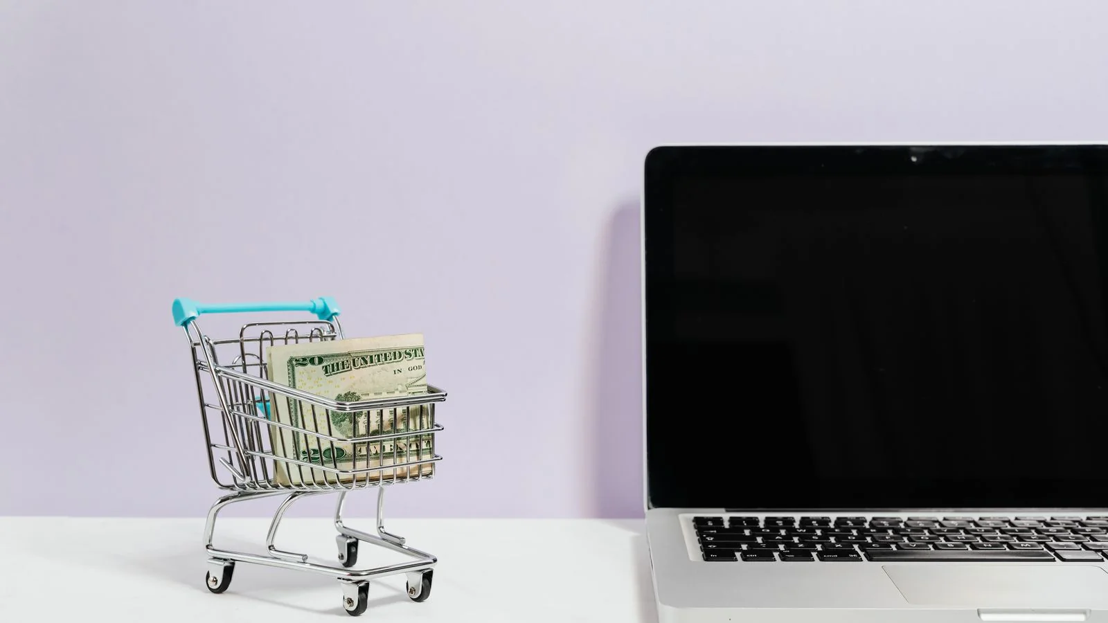 Miniature shopping cart with cash beside a laptop, a buyer weighing what to spend