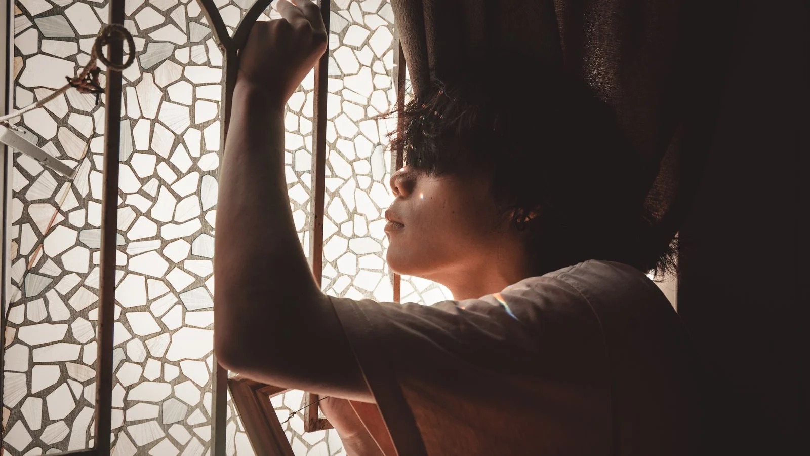A young man standing in thought beside a mosaic glass window in soft light, suggesting the work of forming a view