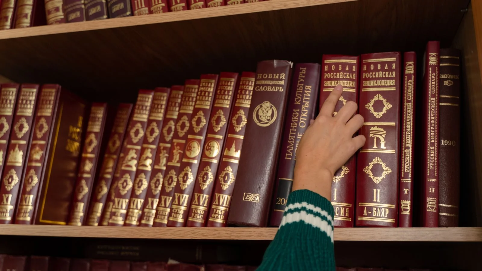A person reaching for a red reference volume on an organised library shelf, the search for independent sources
