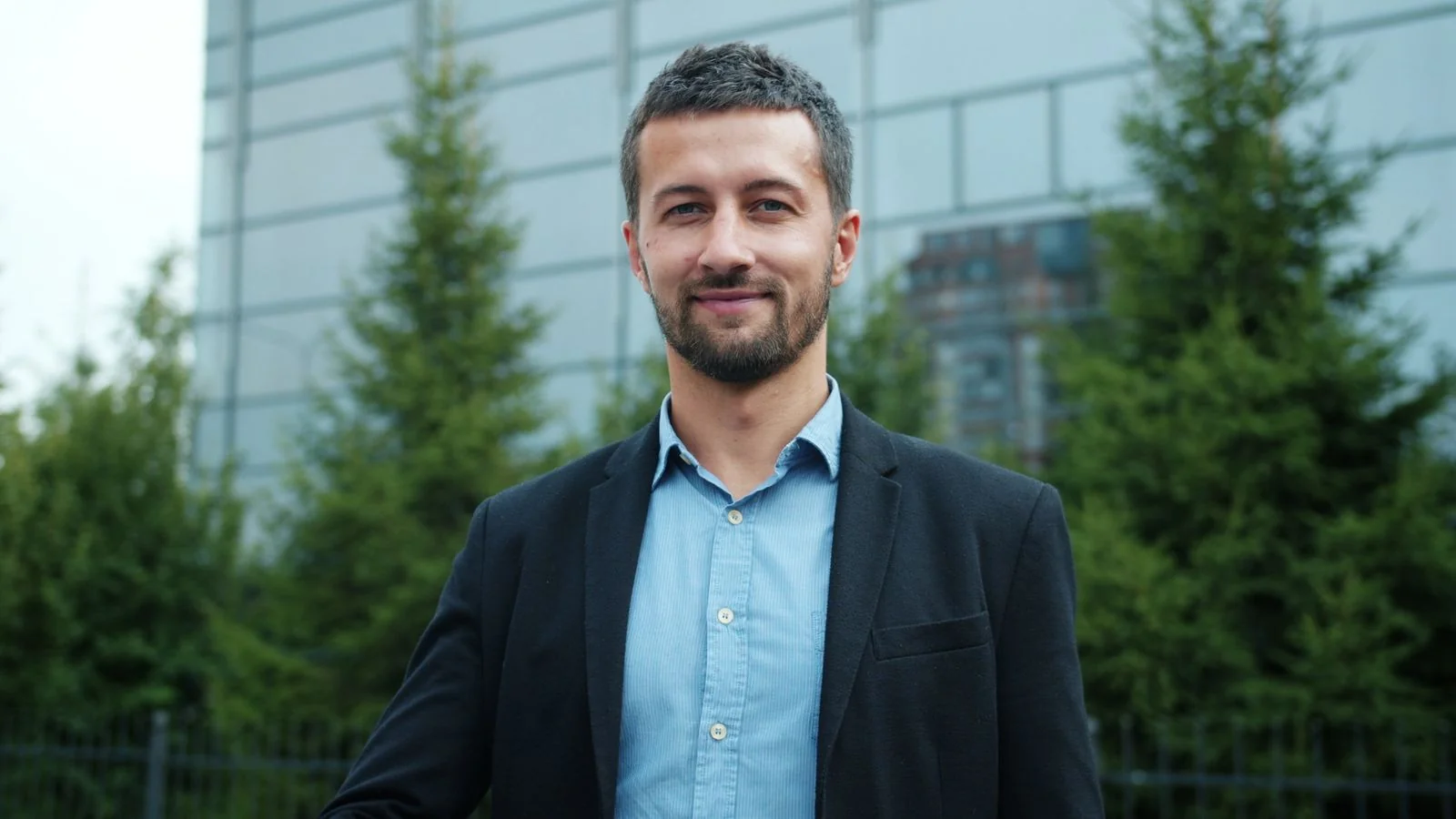 A professional in a blazer standing outdoors in an urban setting, projecting quiet confidence