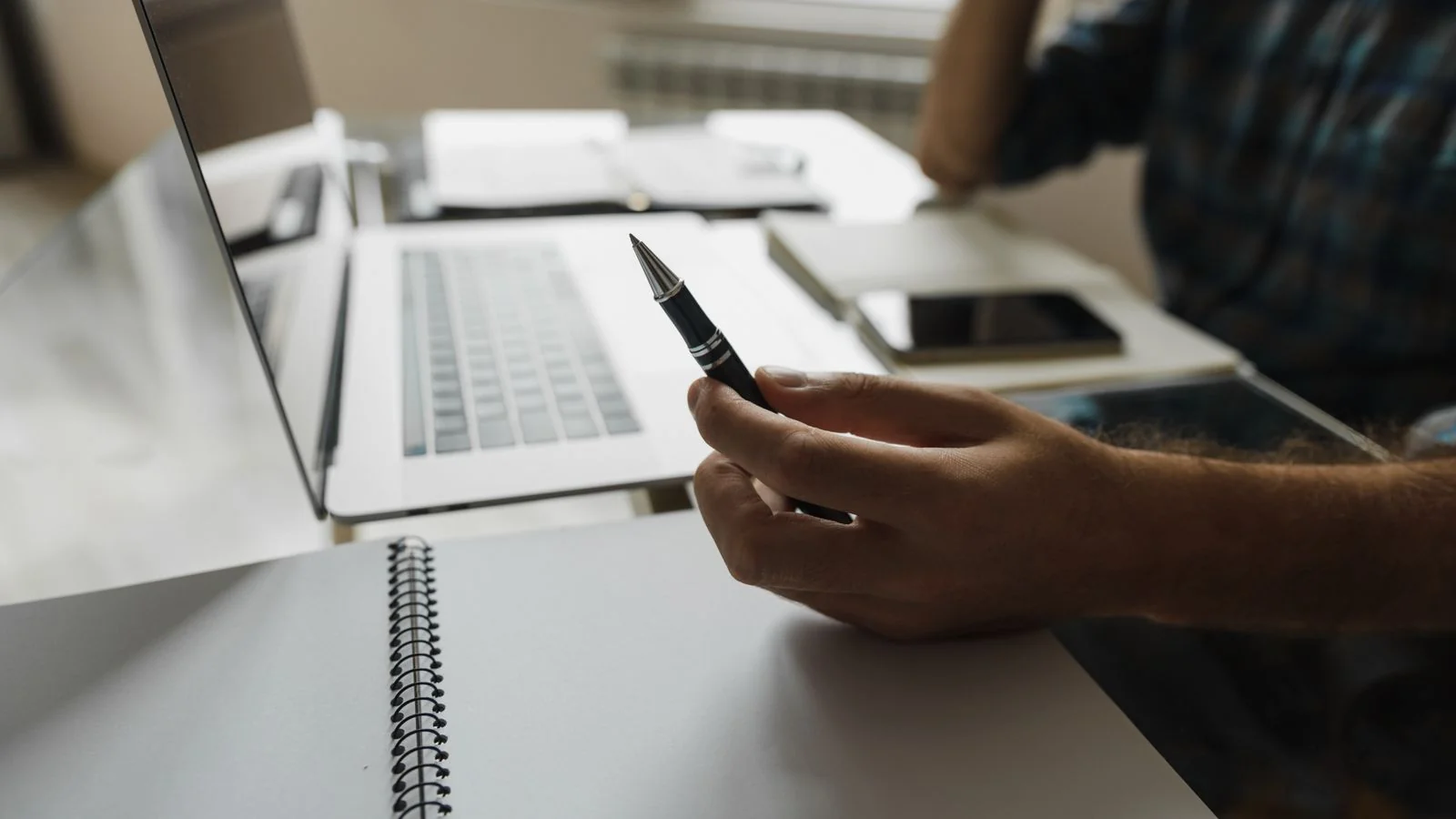 A hand holding a pen beside a laptop and notebook on an office desk
