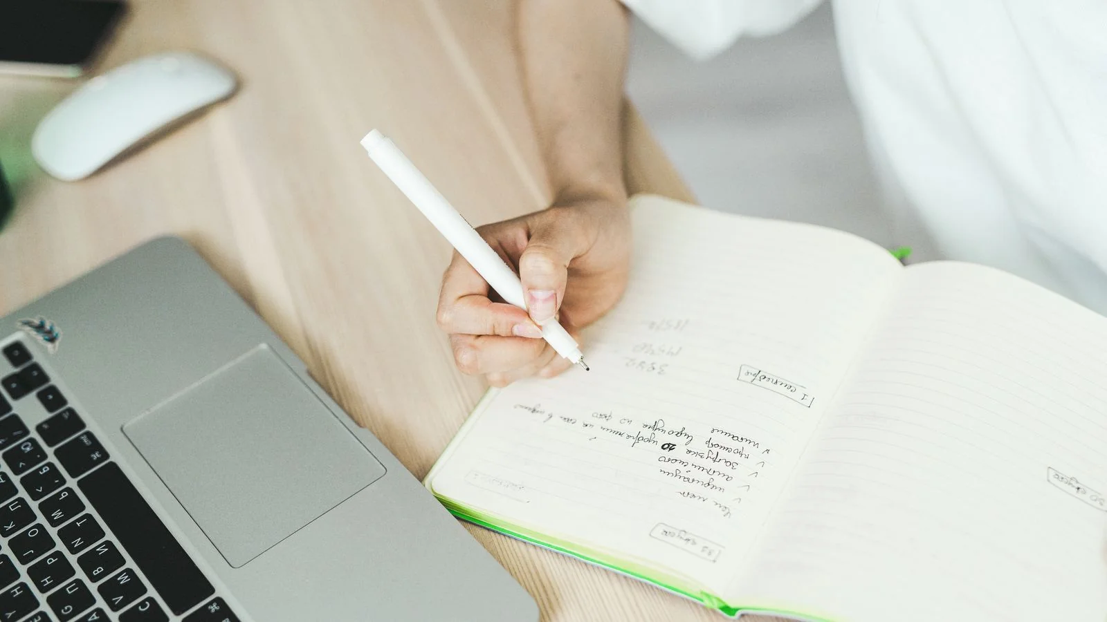 A hand writes in a notebook beside a laptop during an early planning session.