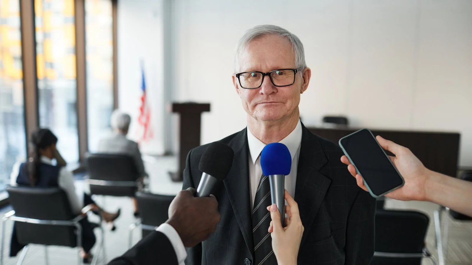 A senior executive being interviewed by a journalist in a conference room, with reporters and broadcast camera operators capturing the response.