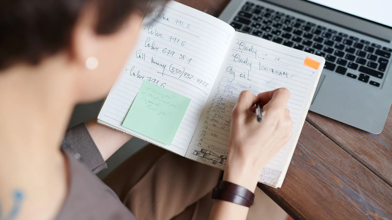 A person writing in a notebook beside a laptop, drafting the opening line that decides everything