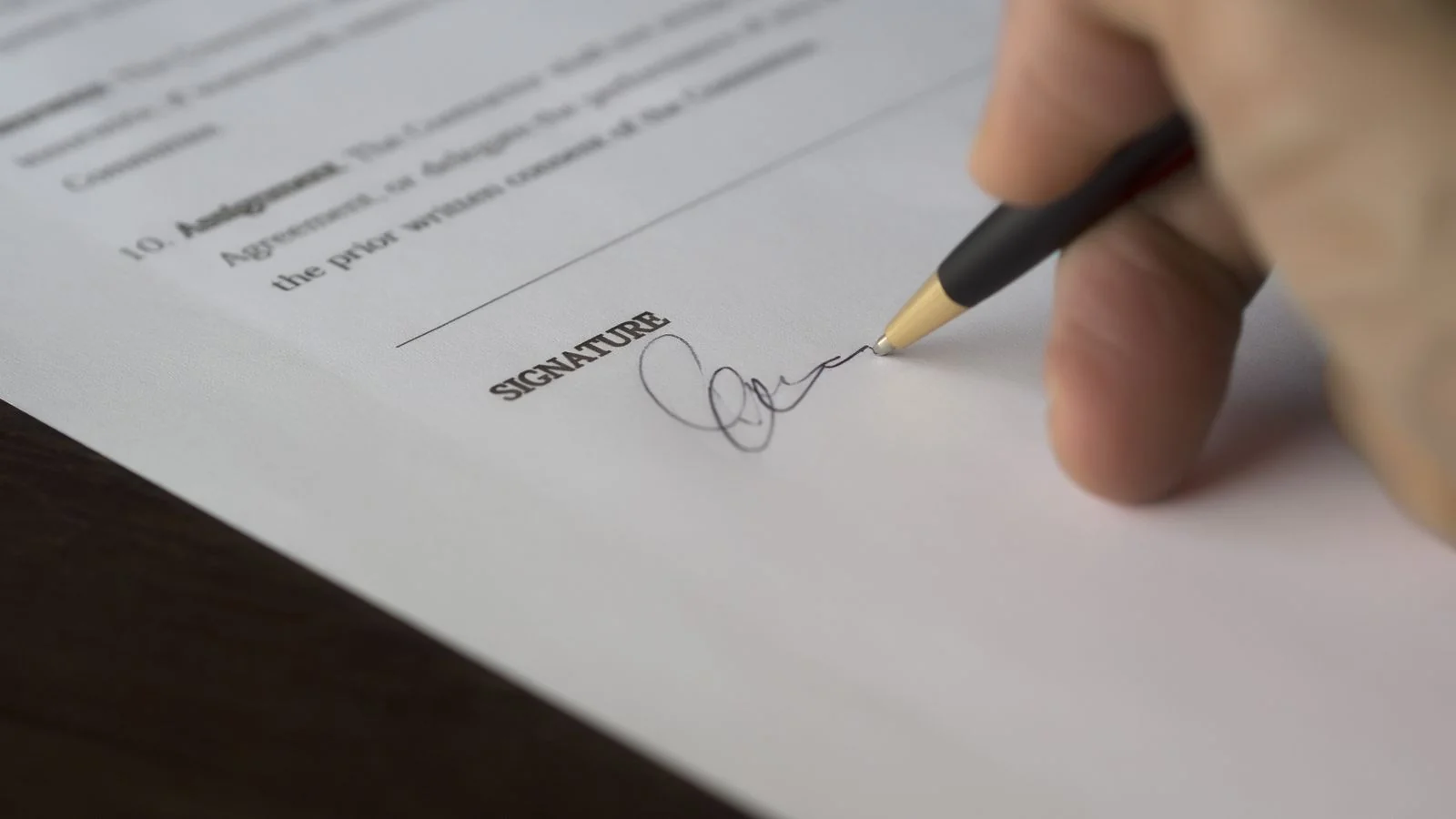 A hand signing a formal document with a pen on a wooden desk