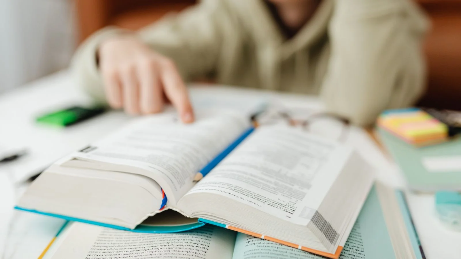 Person seated at a desk pointing to a line of text in an open book, checking a source against a claim