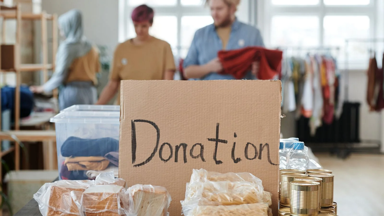 Volunteers sorting donated clothing and food at a donation center beneath a handwritten sign