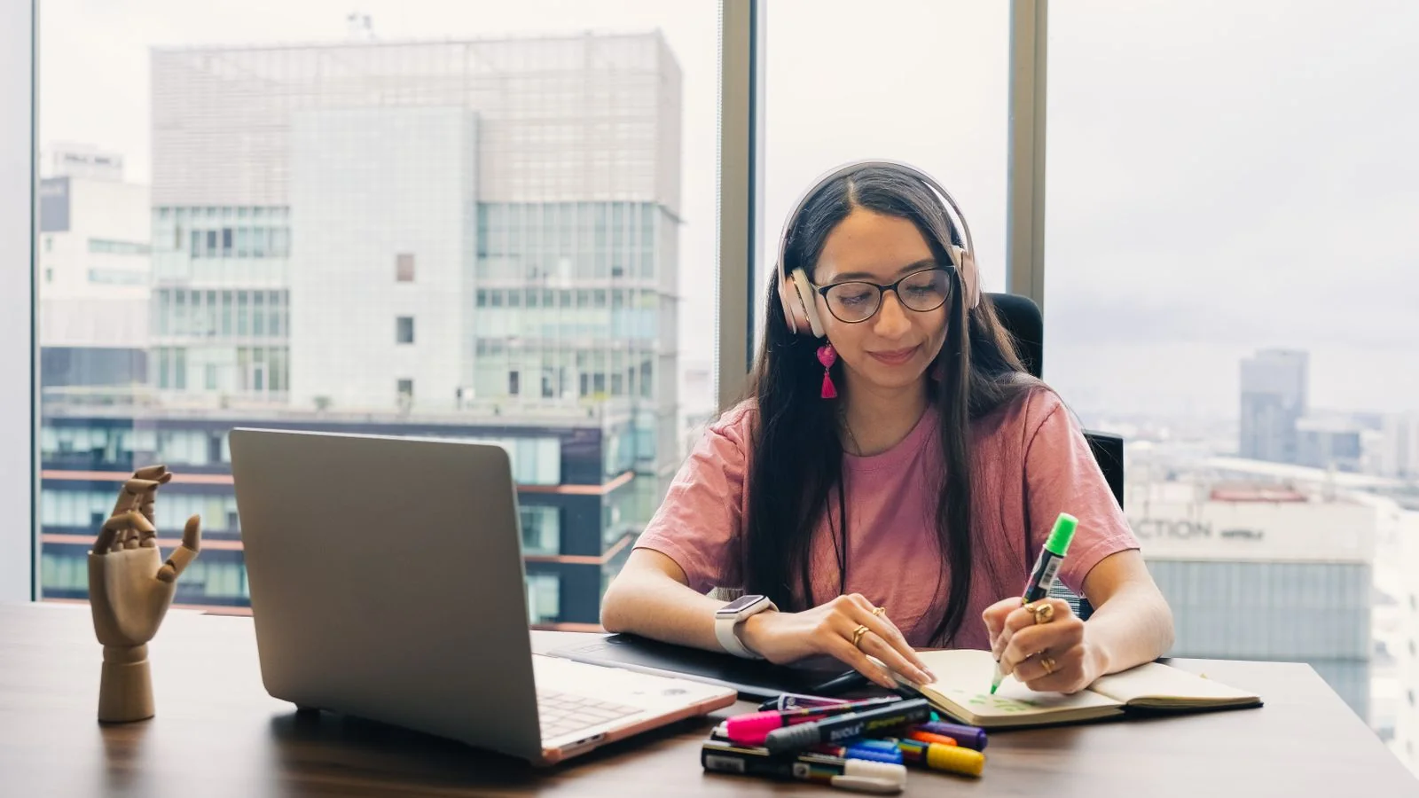 Young woman in headphones writing in a notebook at an office desk with a city view, drafting an op-ed