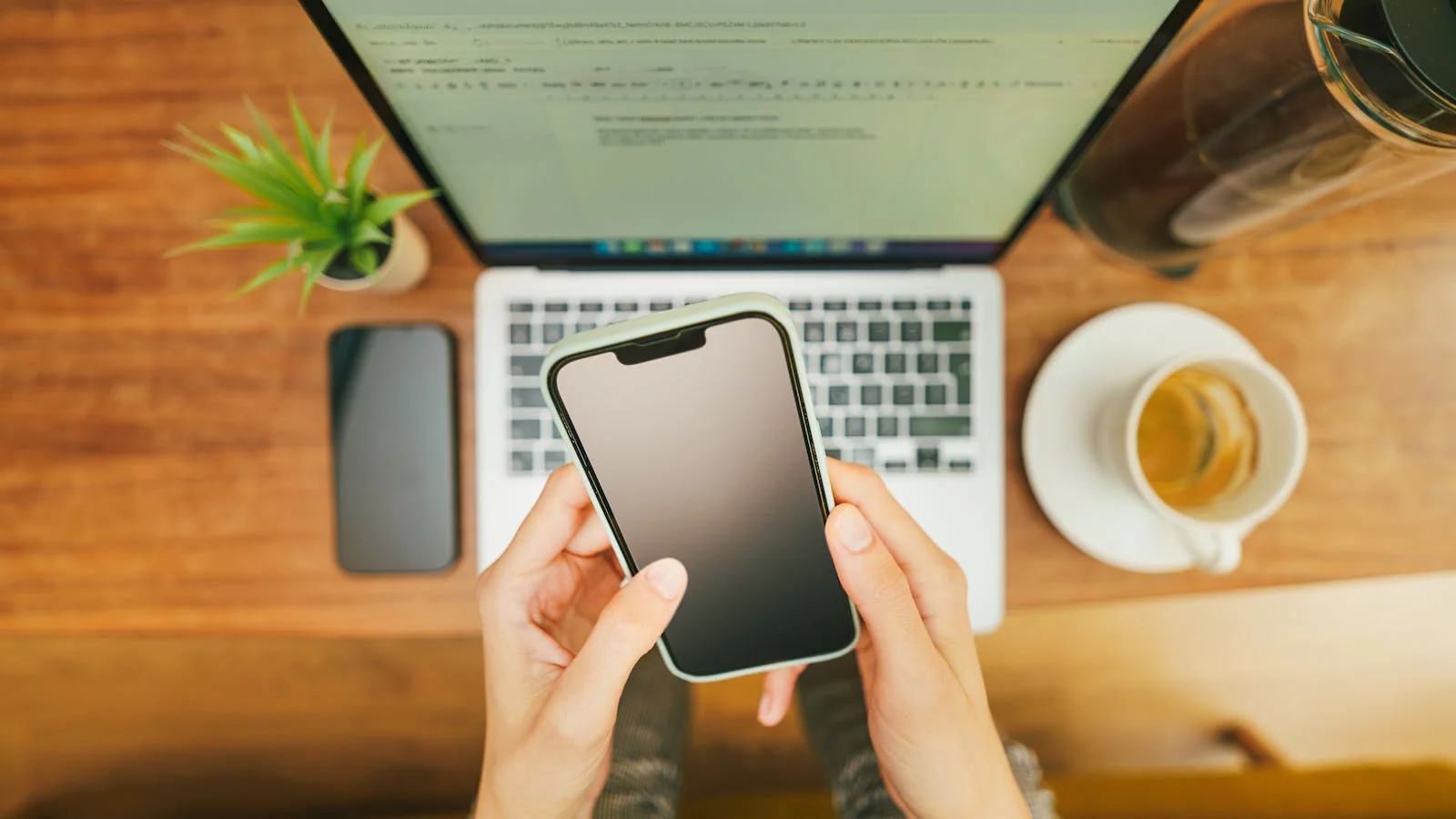 Hands holding a phone above a laptop on a tidy desk, checking an inbox