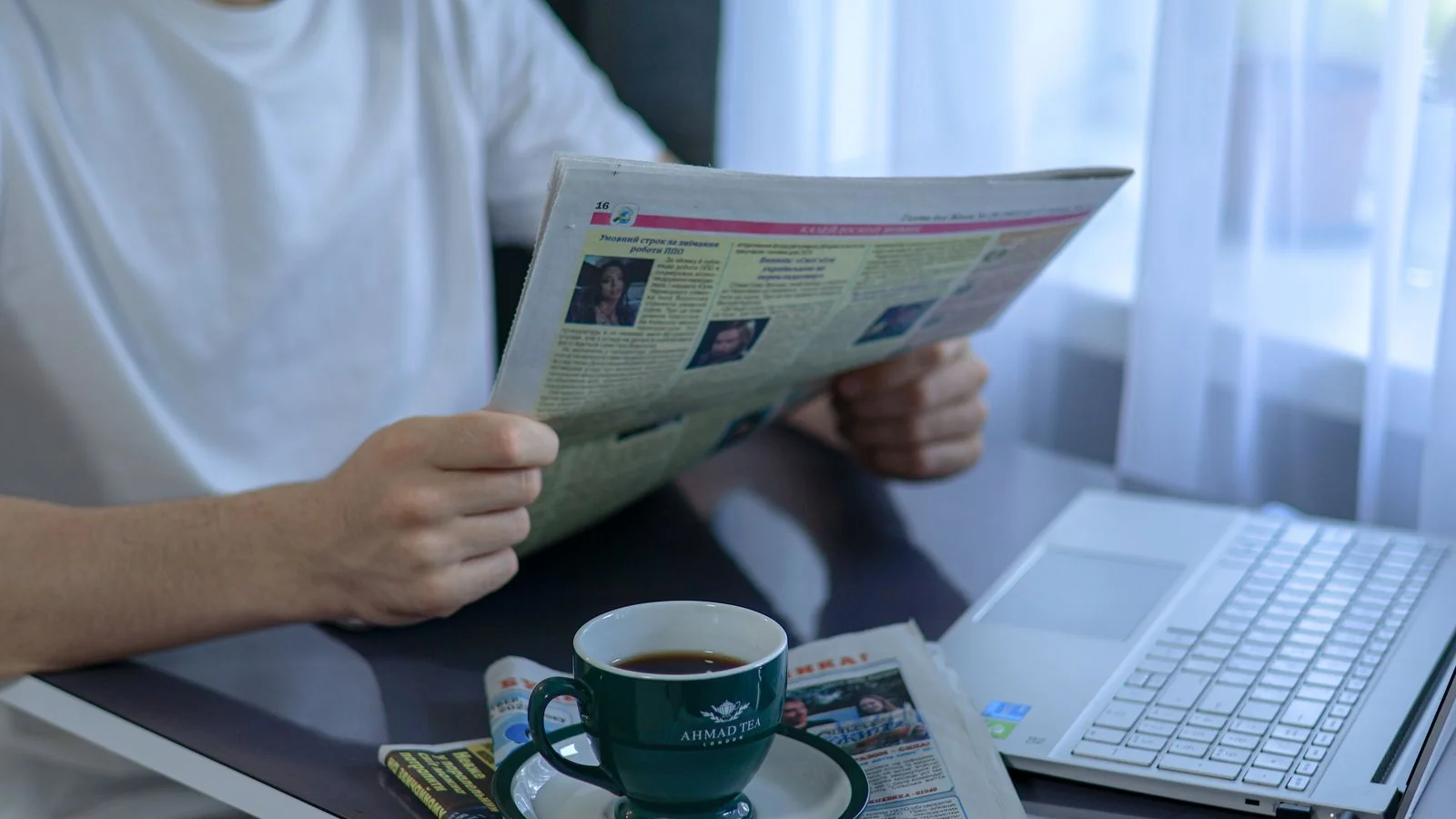 Person reading the newspaper with coffee beside a laptop, catching up on entertainment news