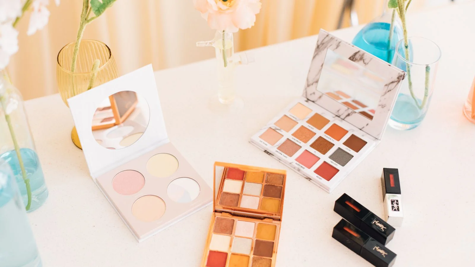 Makeup palettes and lipsticks arranged among flowers on a tabletop