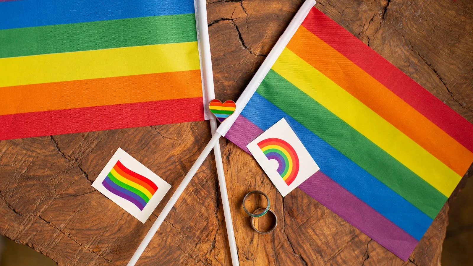 Stacked LGBTQ+ pride flags and community symbols arranged on a wooden surface representing visibility in media