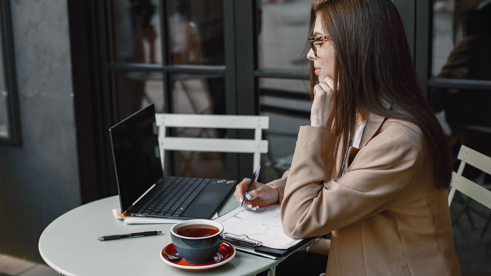 Woman in a brown blazer writing in a notebook beside a laptop at a cafe