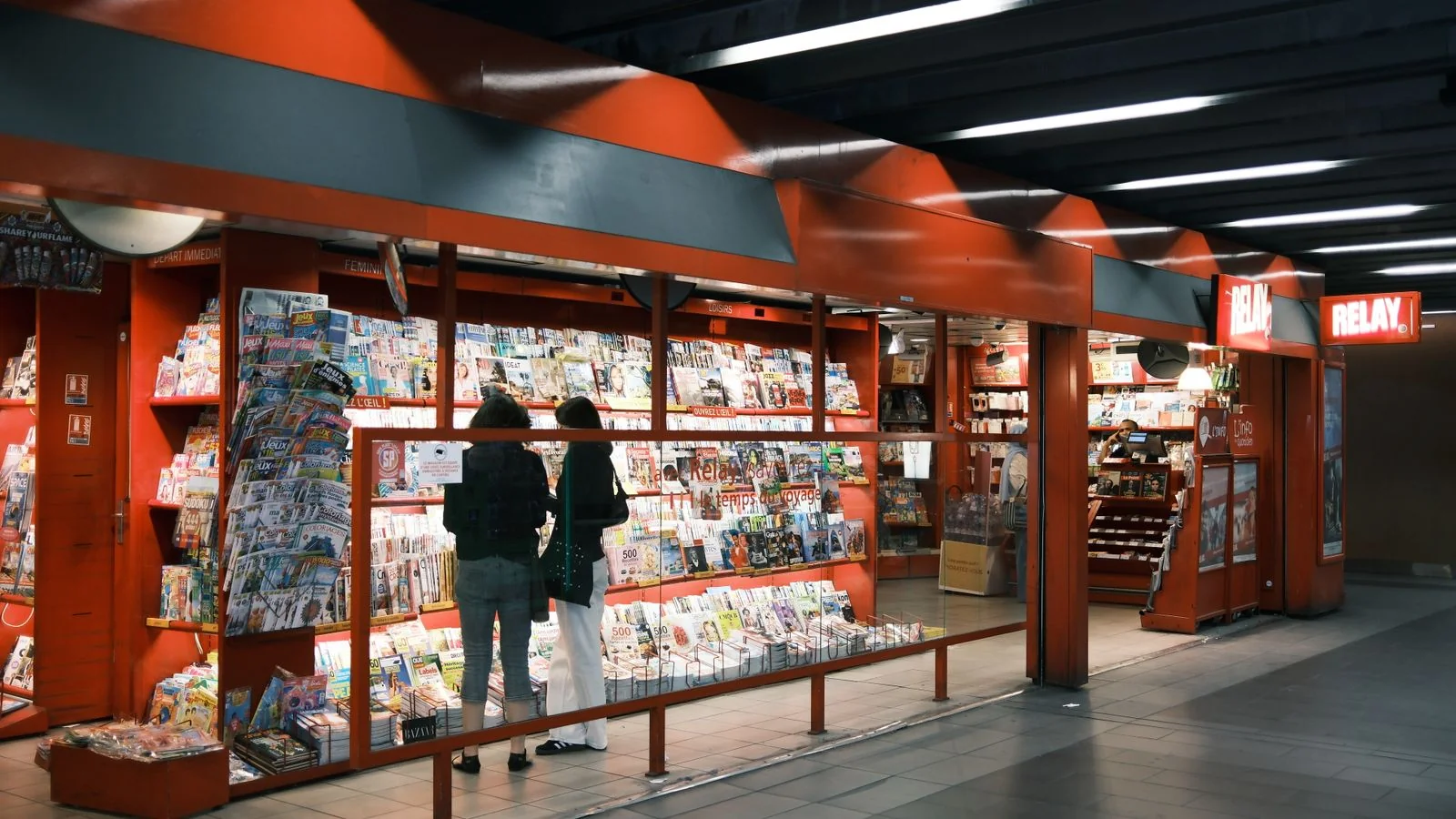 Two adults browsing a newsstand stocked with magazines, where Us Weekly competes for attention