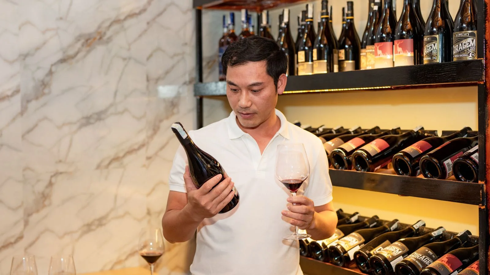 Man in a wine cellar holding a bottle and glass, weighing which wine to pour