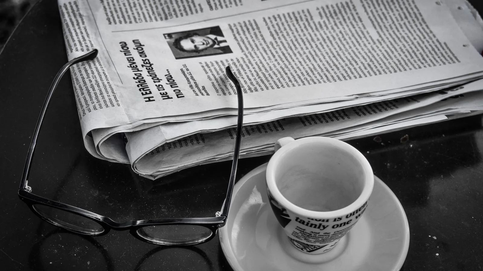 Black and white photo of a newspaper, coffee cup and glasses on a table