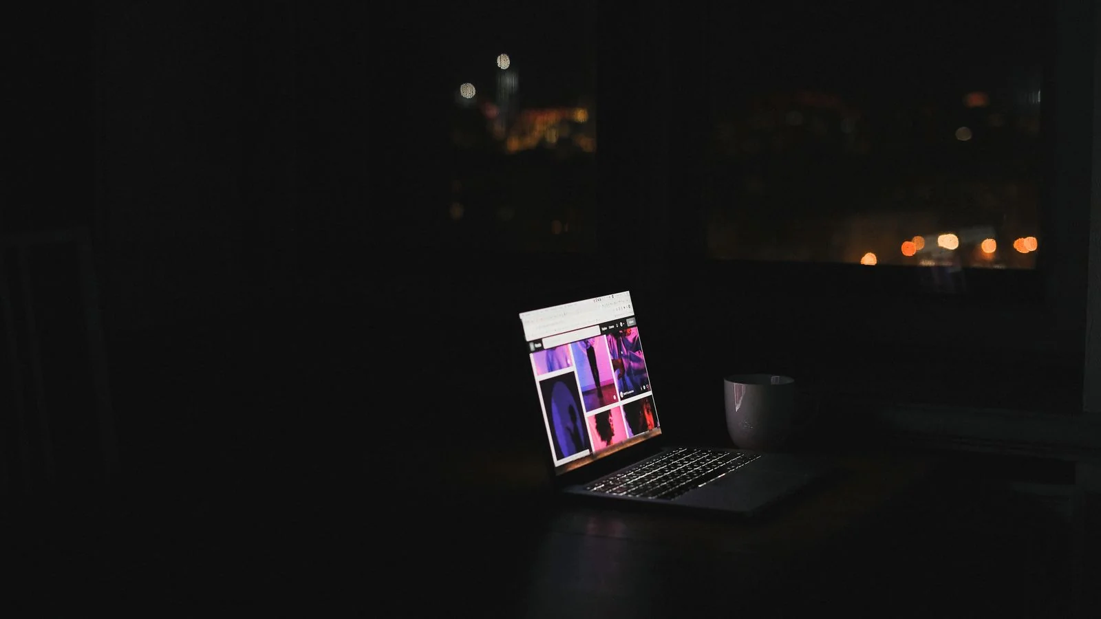 Laptop glowing in a dark room with a city skyline through the window, the late-night submission most contributors make