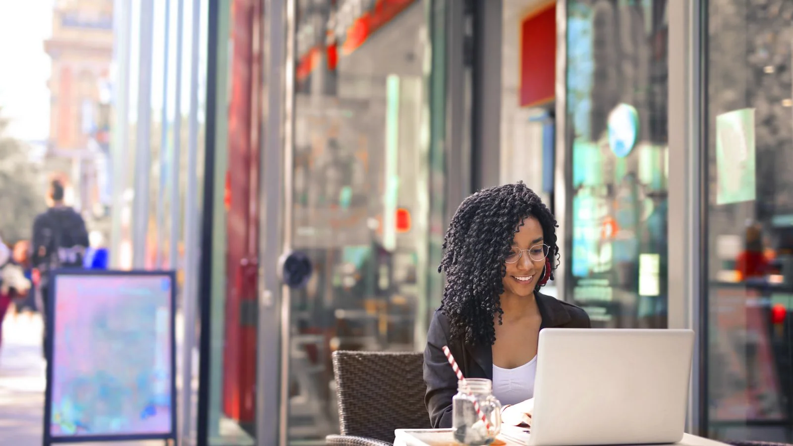 Smiling woman typing on a laptop outside a modern cafe, drafting an expert column for a tech audience