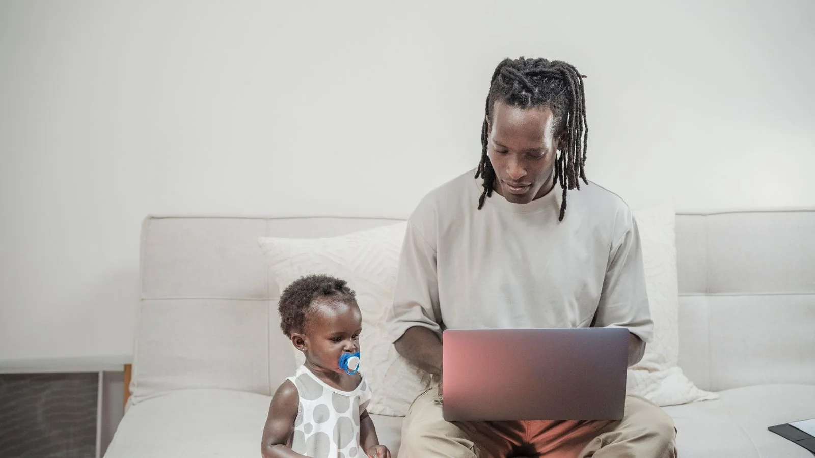 Father working on a laptop at home while his toddler daughter sits beside him