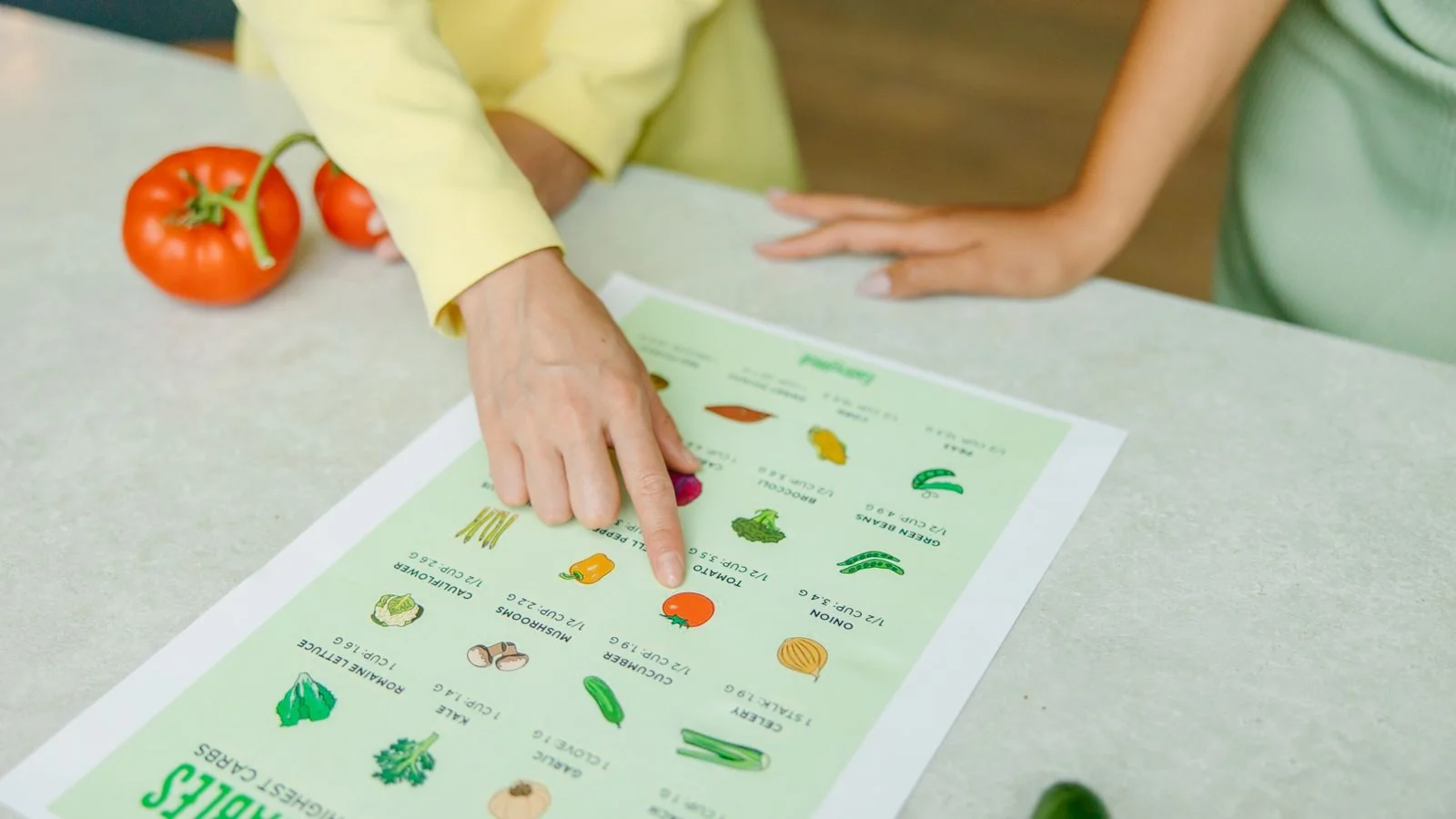 Hands pointing at a vegetable nutrition chart next to fresh tomatoes on a table