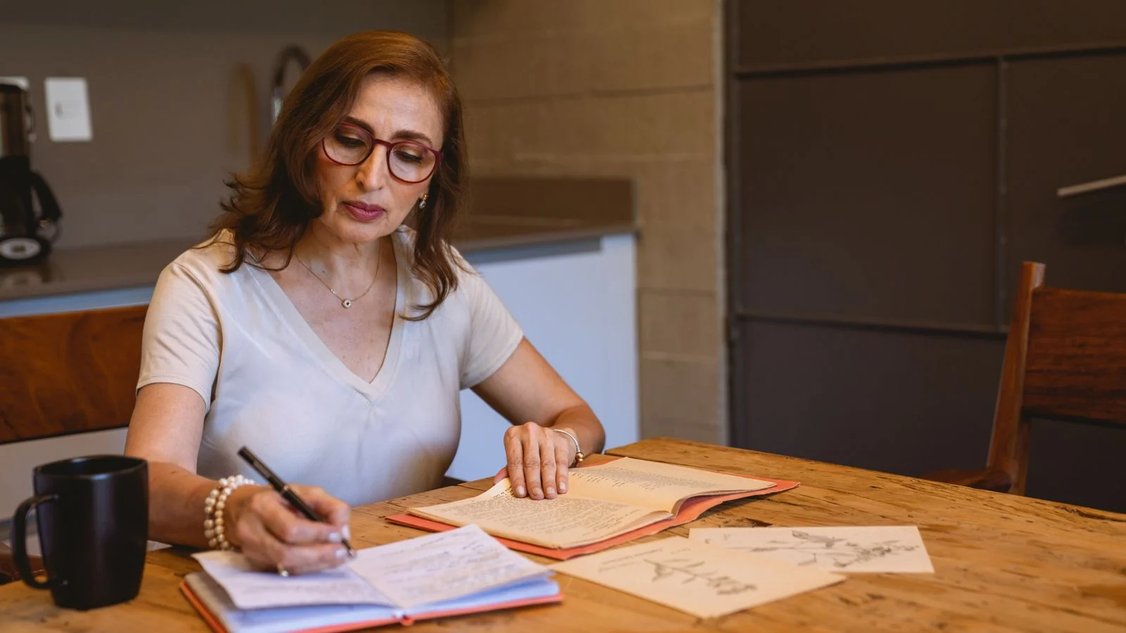 Woman writing notes at a kitchen table with books and coffee, drafting a personal essay