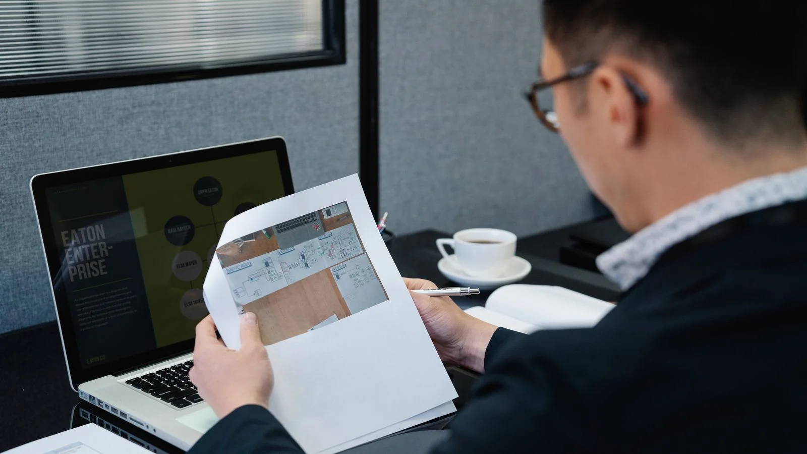 Man reviewing printed documents at an office desk with a laptop open beside him, doing the unglamorous groundwork