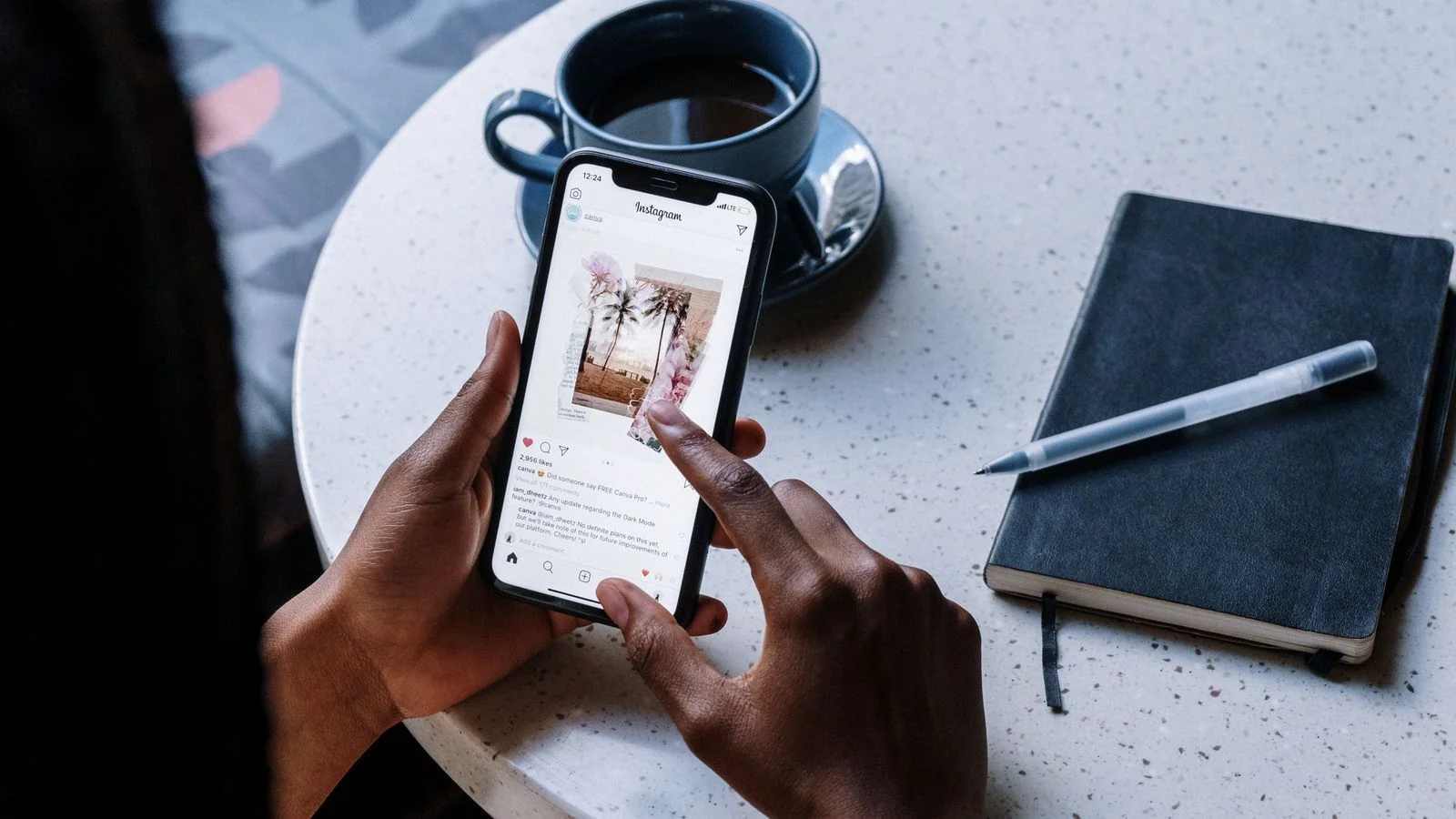 Hands holding a smartphone over a cafe table, scrolling a feed beside a notebook and coffee