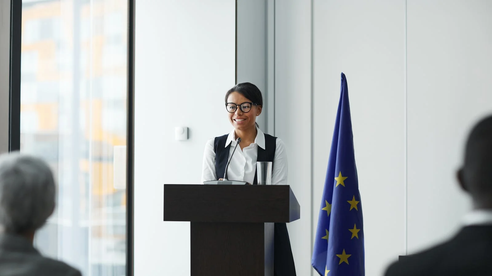 Woman in a sharp blazer giving a confident speech at a conference podium with a national flag behind her