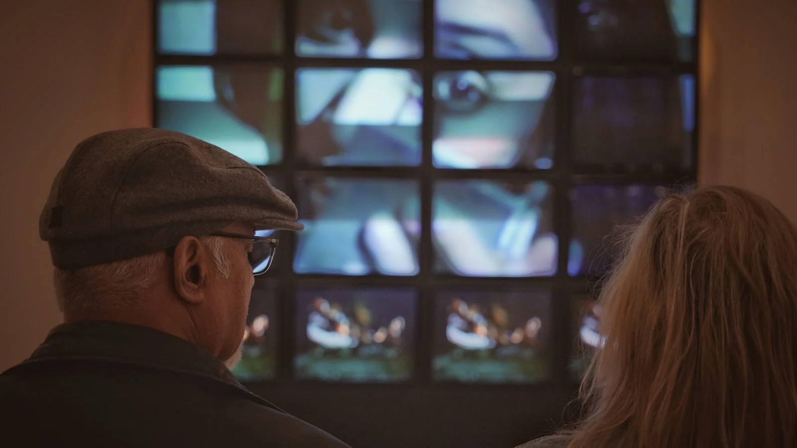 Two gallery visitors studying a multi-screen digital art installation in a darkened room