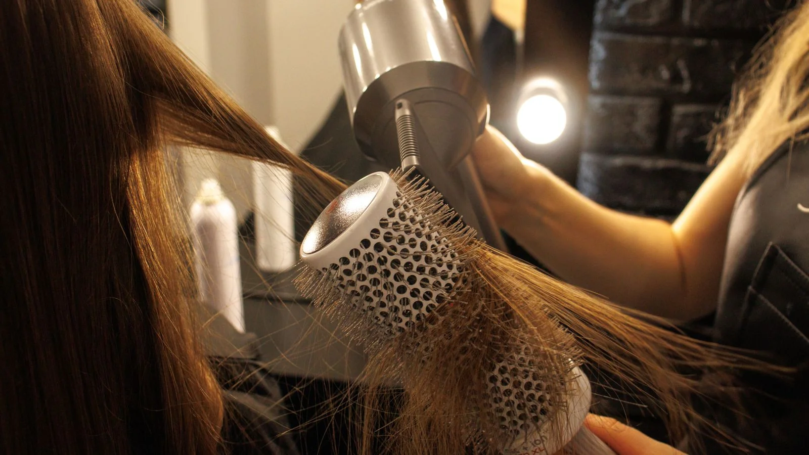 A close-up of a round brush and dryer shaping long hair, the peak-season service booked into editorial pages months earlier