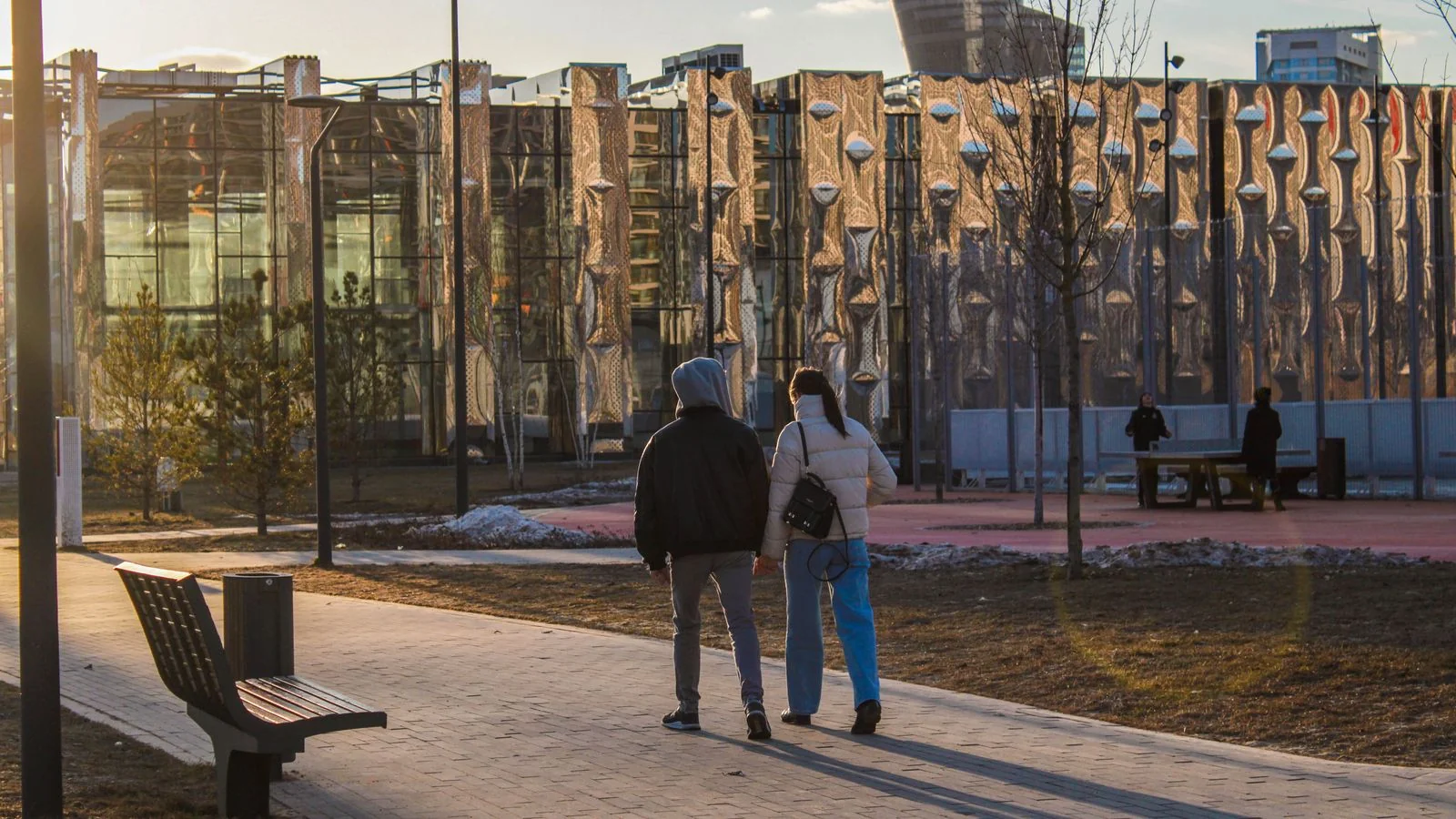 Couple walking through a modern urban park at sunset with contemporary buildings behind them.